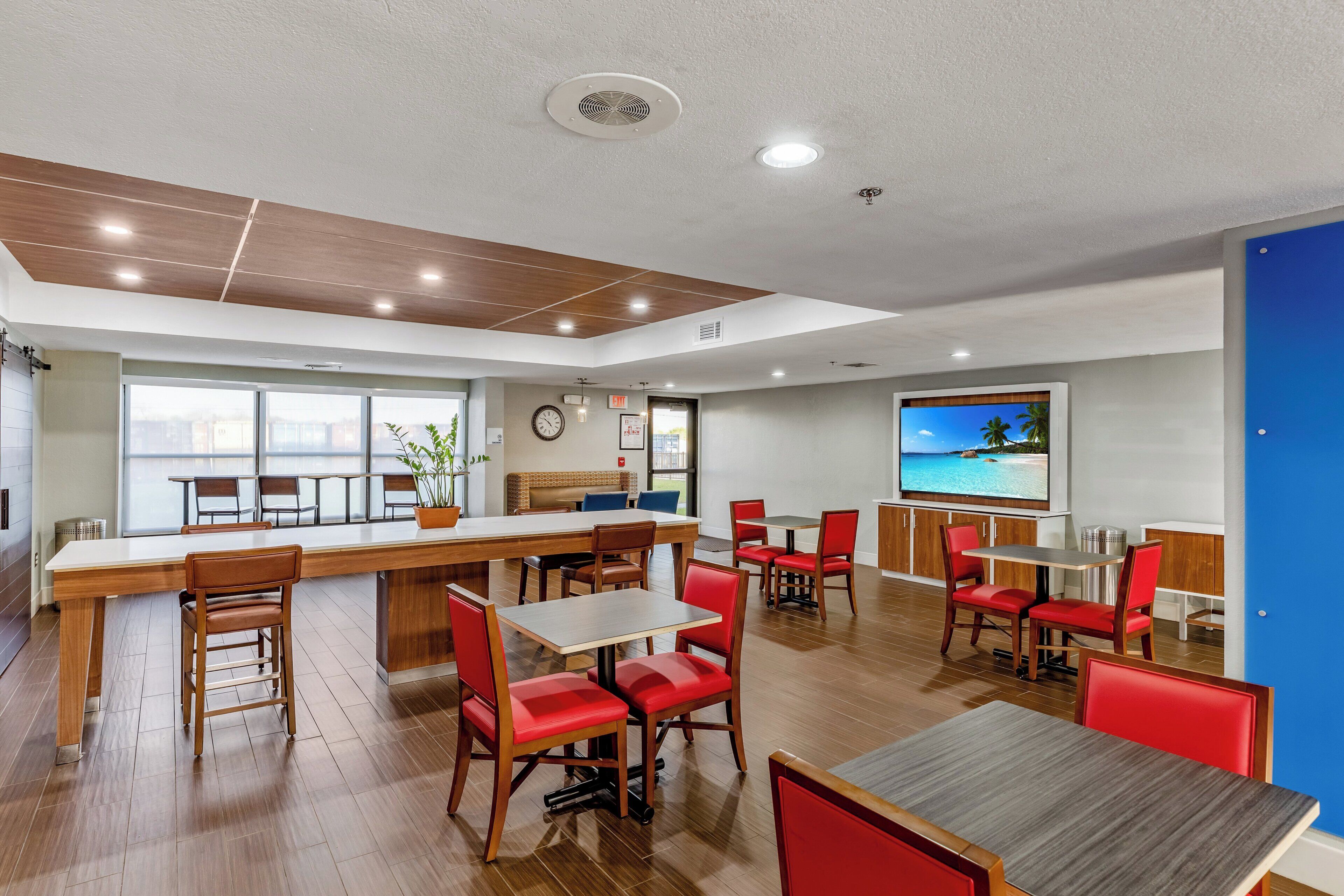A modern dining area with wooden tables and red chairs, a large window with a view, and a clock on the wall.