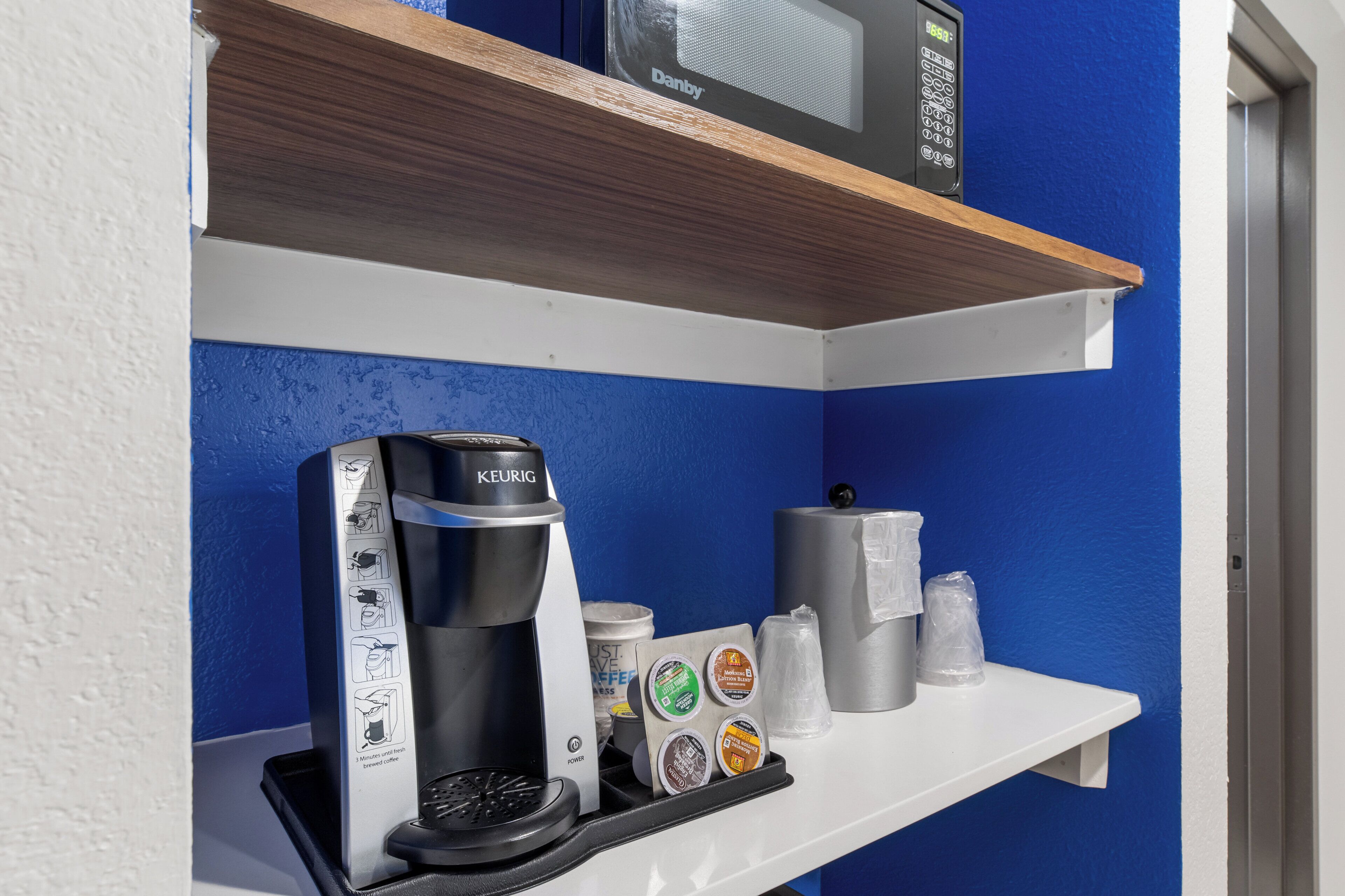 Una máquina de café sobre un estante blanco, con cápsulas de café y un dispensador de toallas de papel.