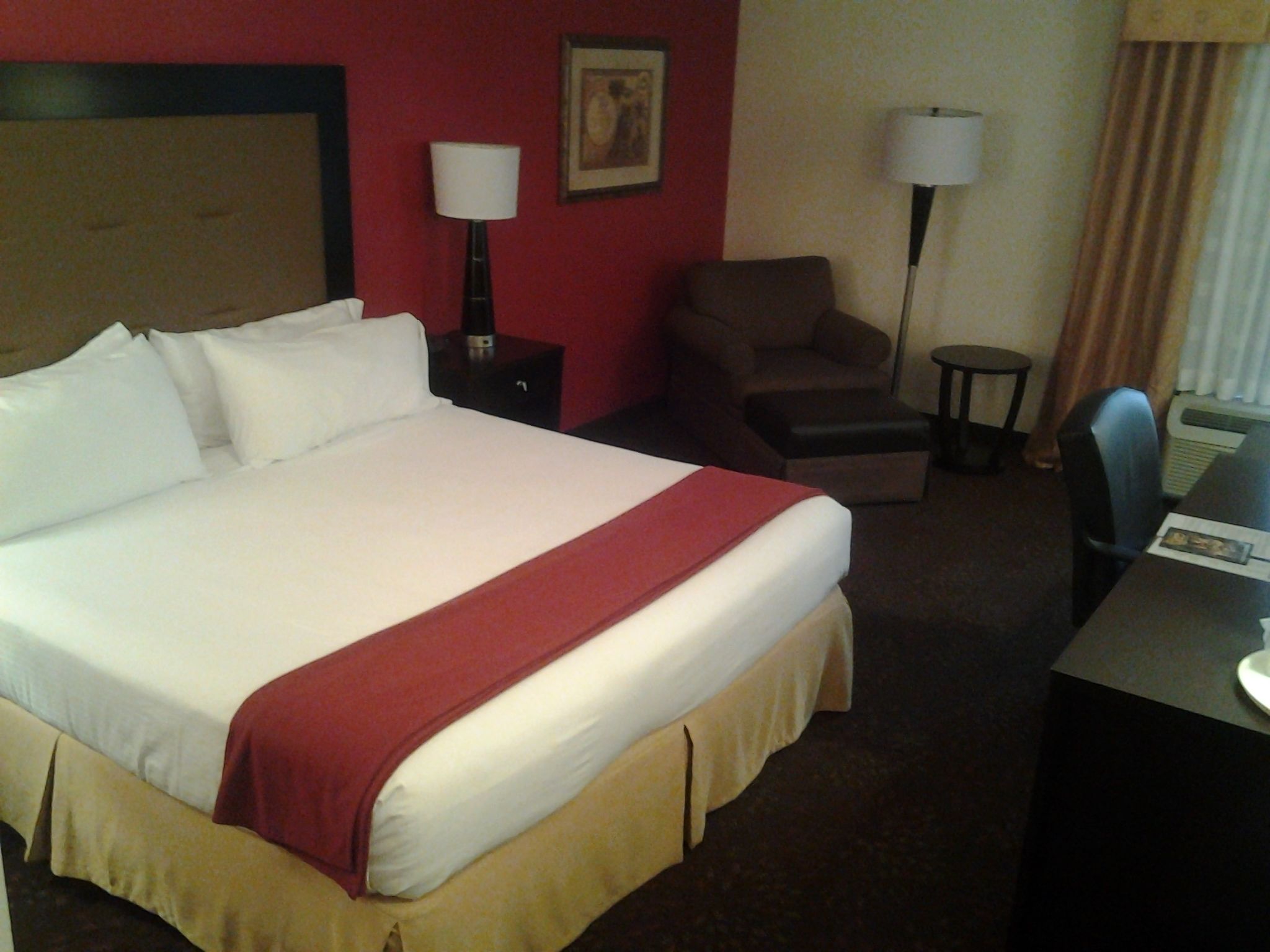 A hotel room with a large bed, a desk, two chairs, a nightstand, and a lamp.