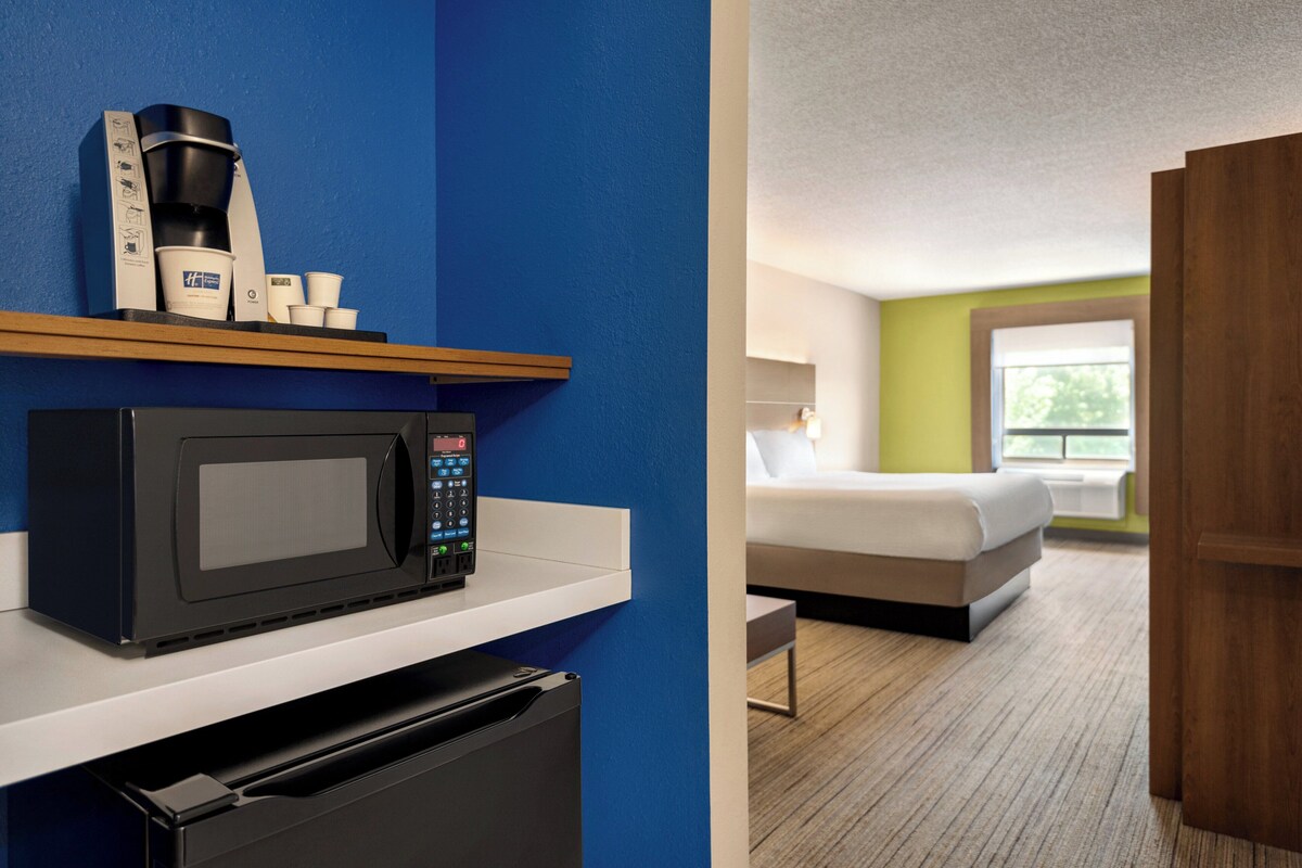 Quarto de hotel com micro-ondas, cafeteira e chávenas numa prateleira, cama com vista para o exterior, e porta de madeira.