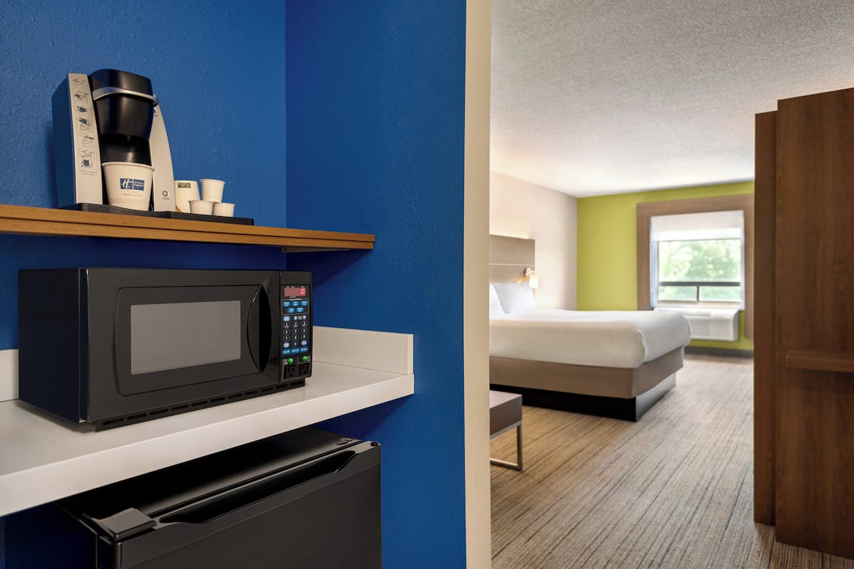 Ein Hotelzimmer mit Mikrowelle, Kaffeemaschine und Tassen auf einem Regal, ein Bett mit Blick ins Freie und eine Holztür.