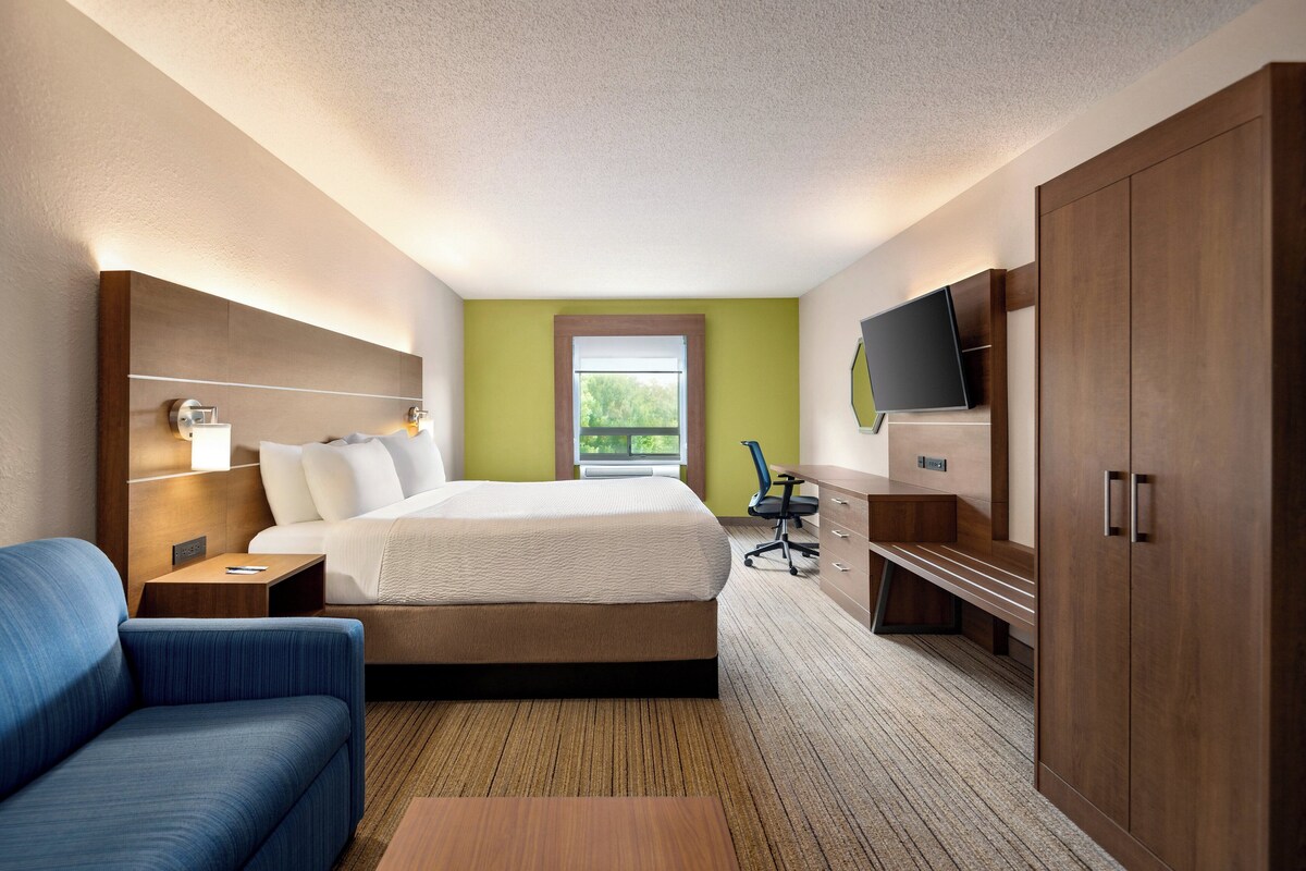 Quarto de hotel com cama, escrivaninha, cadeira, televisão e guarda-roupa.