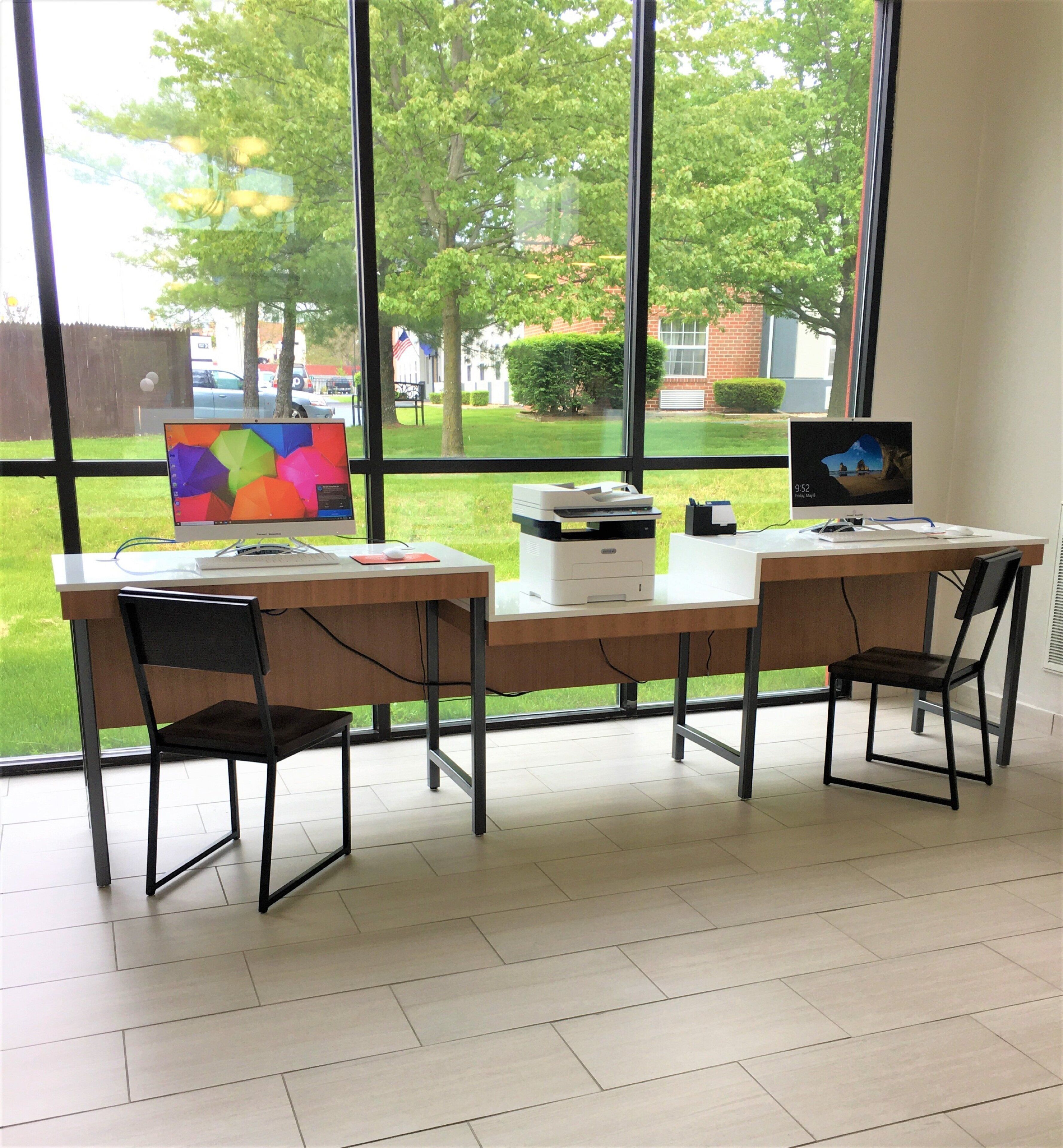 Un espacio de trabajo con dos escritorios, sillas, una computadora, una impresora y una ventana que ofrece vistas a un paisaje verde y edificios.
