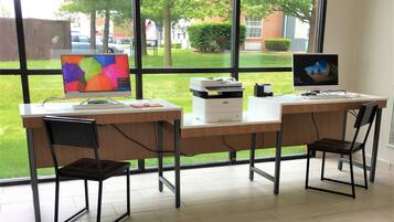 Un espacio de trabajo con dos escritorios, sillas, una computadora, una impresora y una ventana que ofrece vistas a un paisaje verde y edificios.