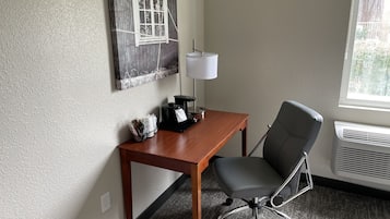 Ett hotellrum med ett skrivbord i trä, en svart kontorsstol, en kaffebryggare, en lampa och ett fönster med utsikt över träd.