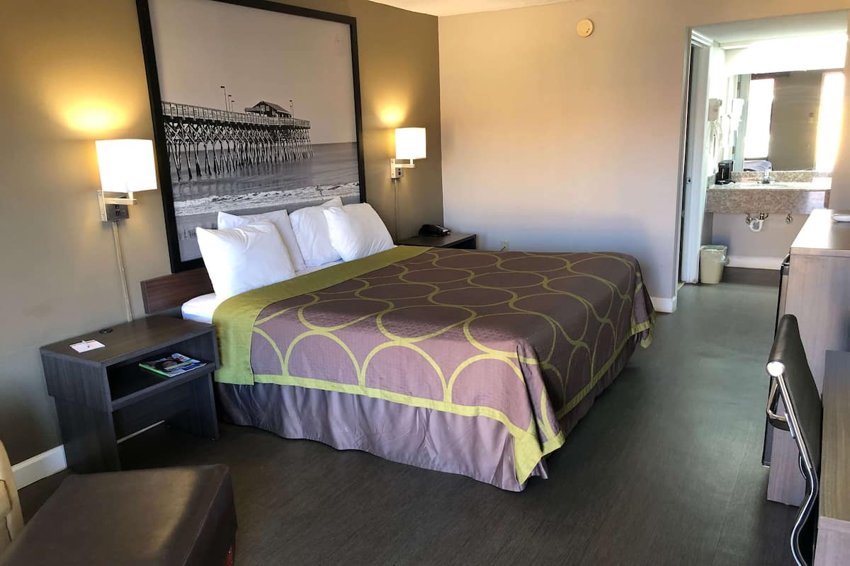 Habitación de hotel con una cama grande, dos lámparas de noche, una mesita de noche, una pequeña mesa, un banco, un televisor montado en la pared y un baño visible a través de una puerta abierta.