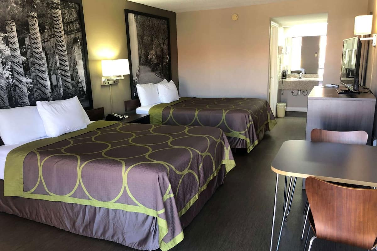 Habitación de hotel con dos camas, un escritorio, una silla, un televisor y un baño visible a través de una puerta abierta.