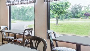 Ein Restaurantinnenraum mit Tischen und Stühlen, große Fenster mit Blick auf eine Straße und Grünflächen.