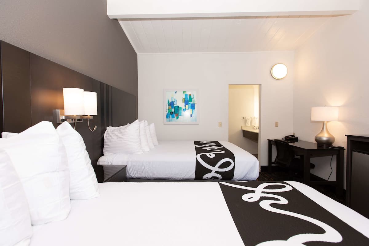 Een hotelkamer met twee bedden, een bureau, een stoel, een lamp en een kunstwerk aan de muur.