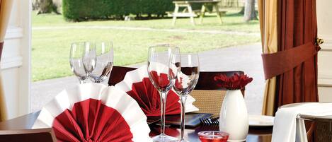 A dining table set for two with wine glasses, a vase with red flowers, and a fan.