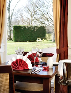 A dining table set for two with wine glasses, a vase with red flowers, and a fan.
