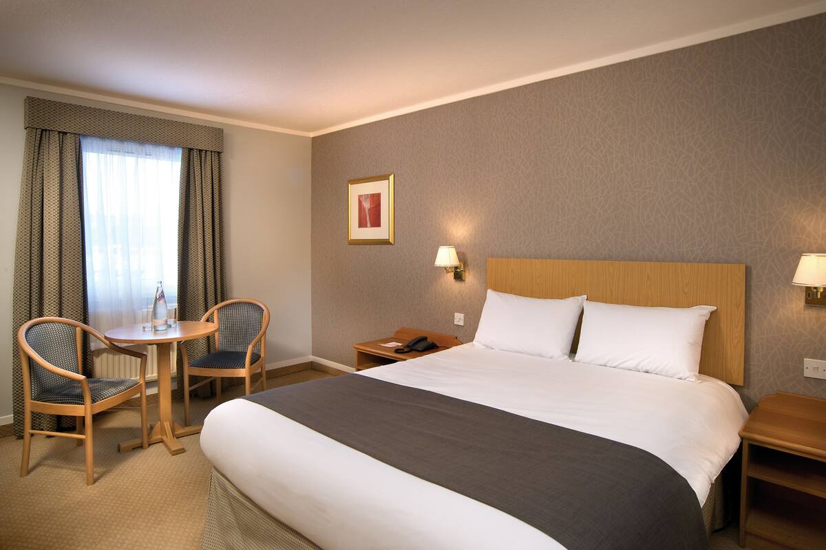A hotel room with a large bed, two chairs, a small table, a nightstand, and a lamp.