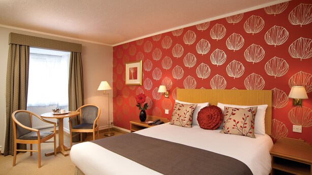 A hotel room with a large bed, two chairs, a small table, a nightstand with a lamp, and a wall with a leaf pattern. - The Hickstead Hotel (Haywards Heath)