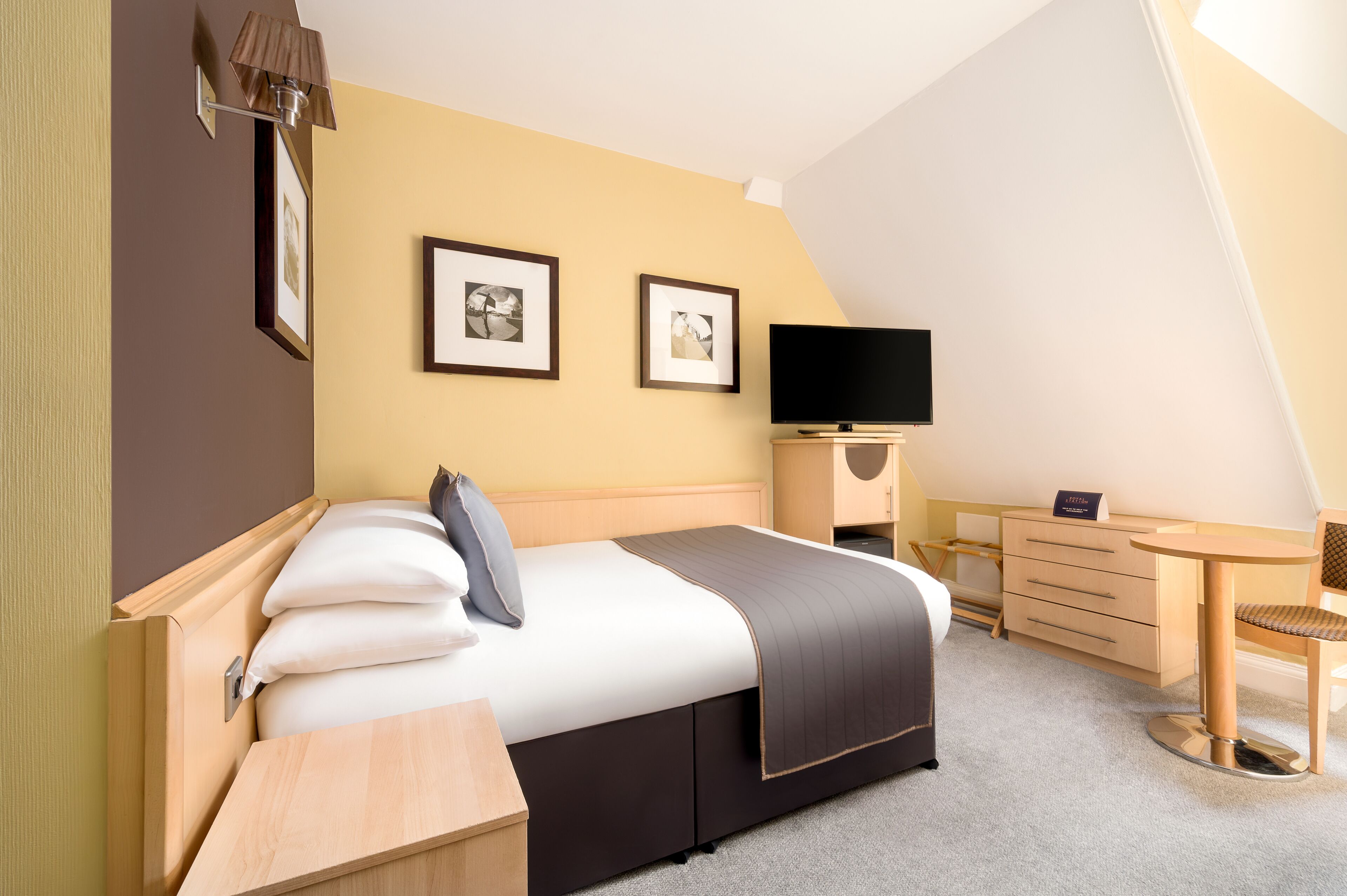 Ein ordentlich eingerichtetes Hotelzimmer mit einem Bett, einem Schreibtisch, einem Stuhl, einem Fernseher und zwei gerahmten Bildern an der Wand.