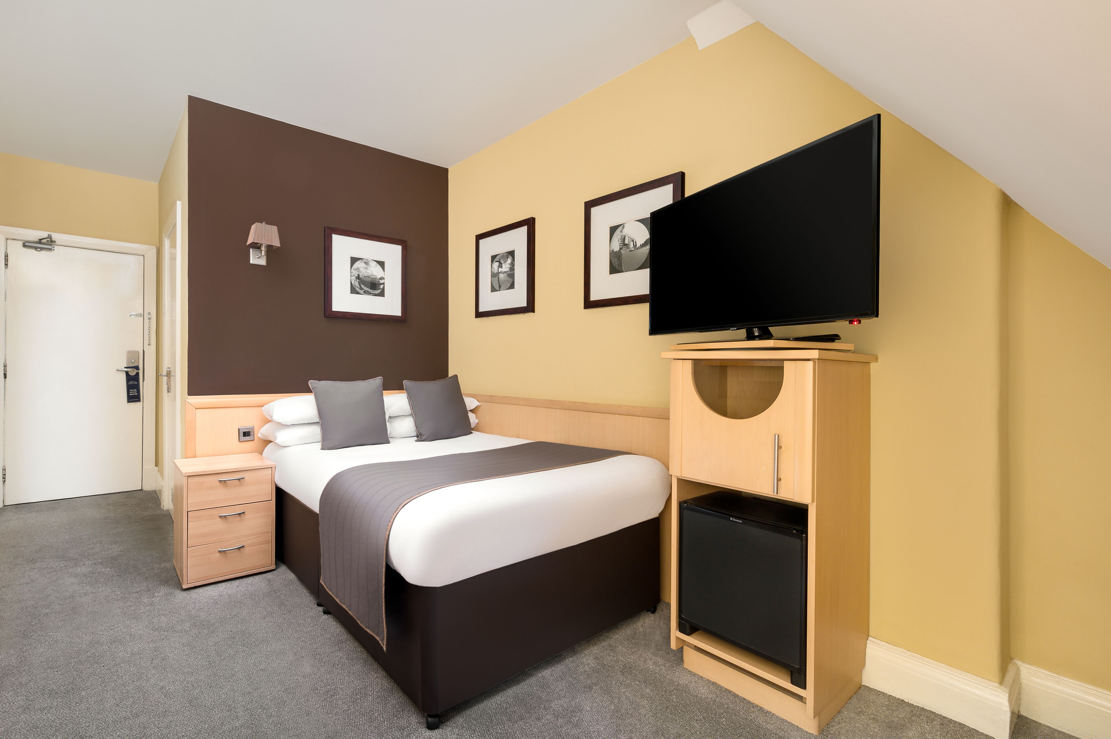 A hotel room with a bed, a TV, a small wooden cabinet, and framed pictures on the wall.