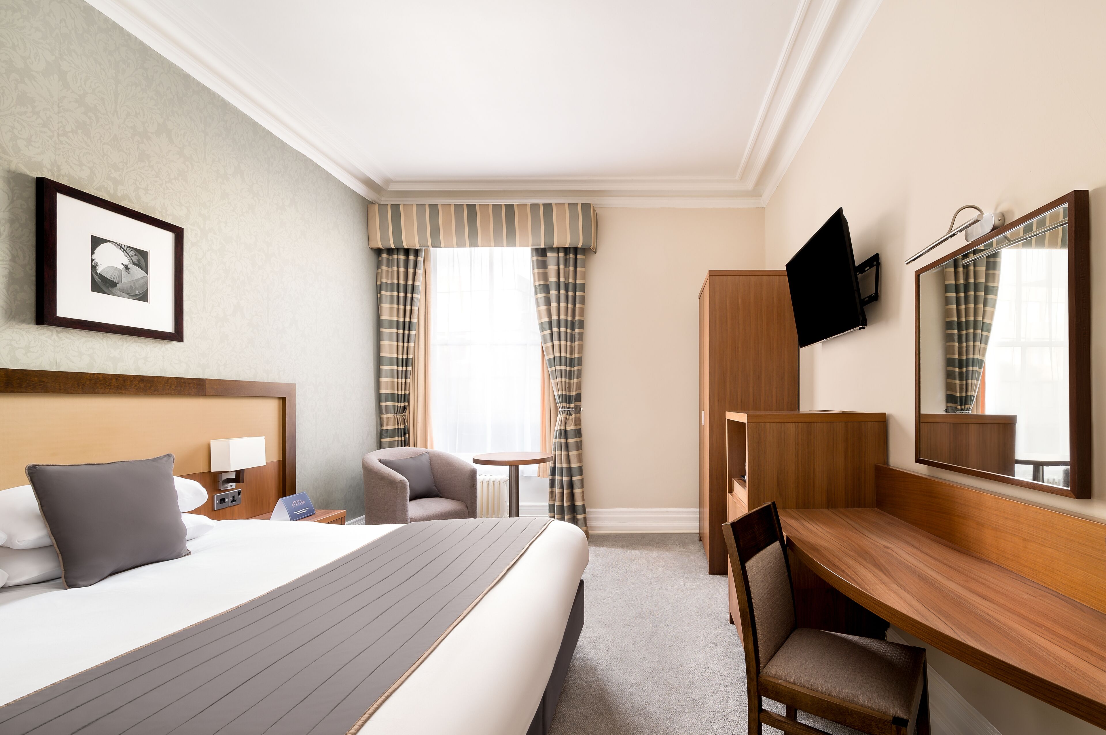 A hotel room with a large bed, a desk, a chair, a small table, a TV, and a mirror.