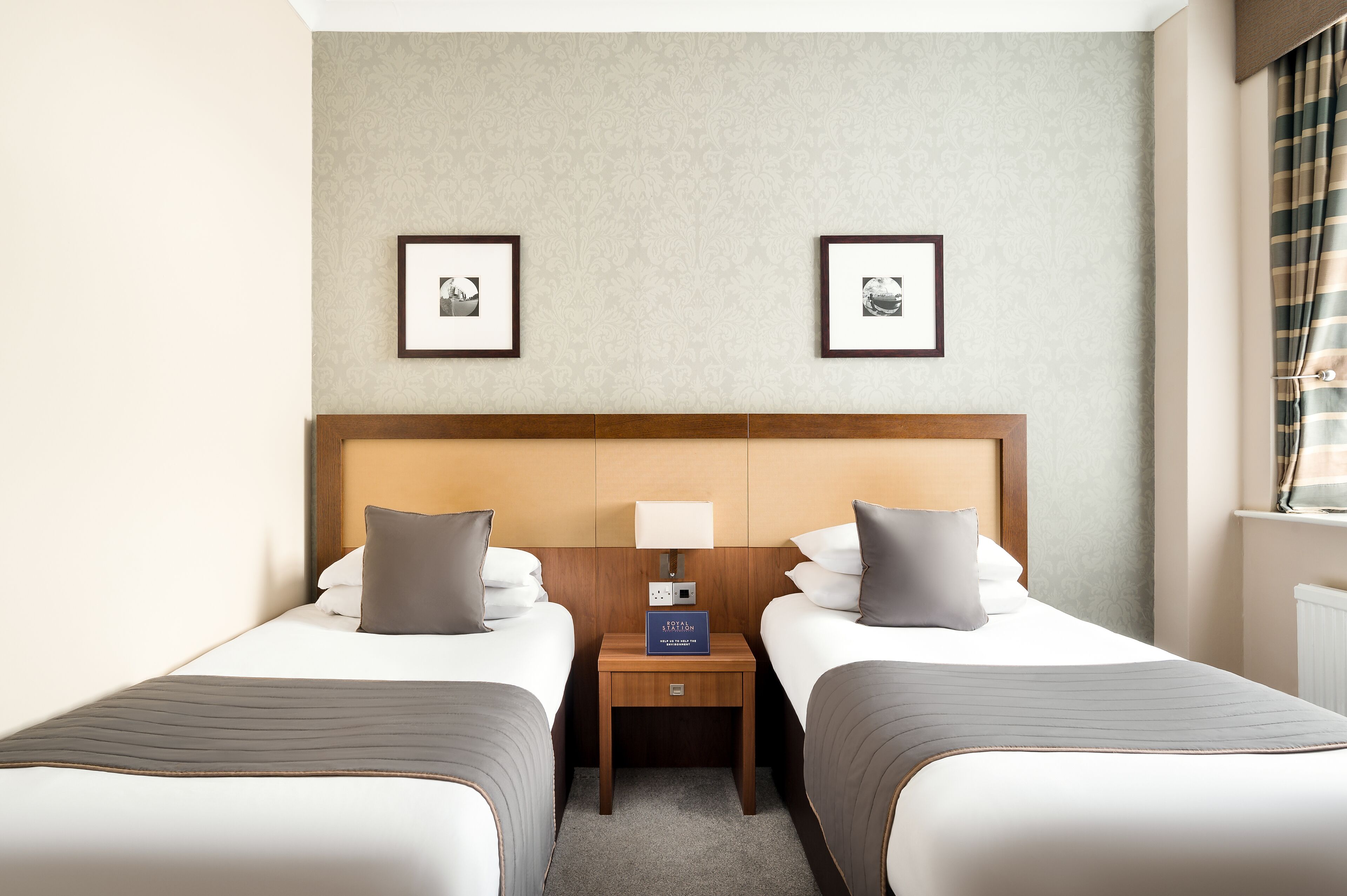 Une chambre d’hôtel avec deux lits, une tête de lit en bois, une table de chevet avec une lampe et des tableaux encadrés au mur.