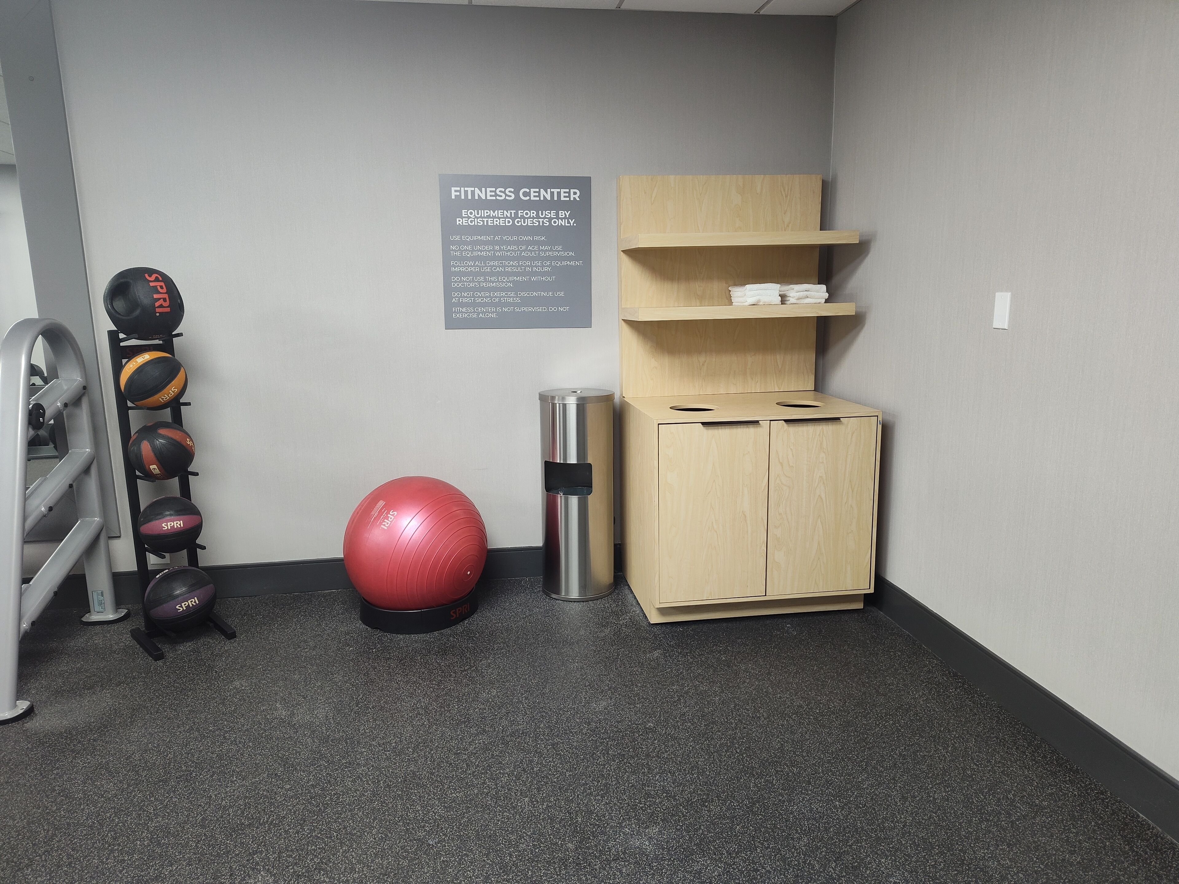 A fitness center with exercise equipment, a red exercise ball, a metal trash can, and a wooden cabinet with shelves.