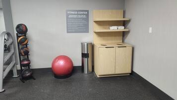 A fitness center with exercise equipment, a red exercise ball, a metal trash can, and a wooden cabinet with shelves.