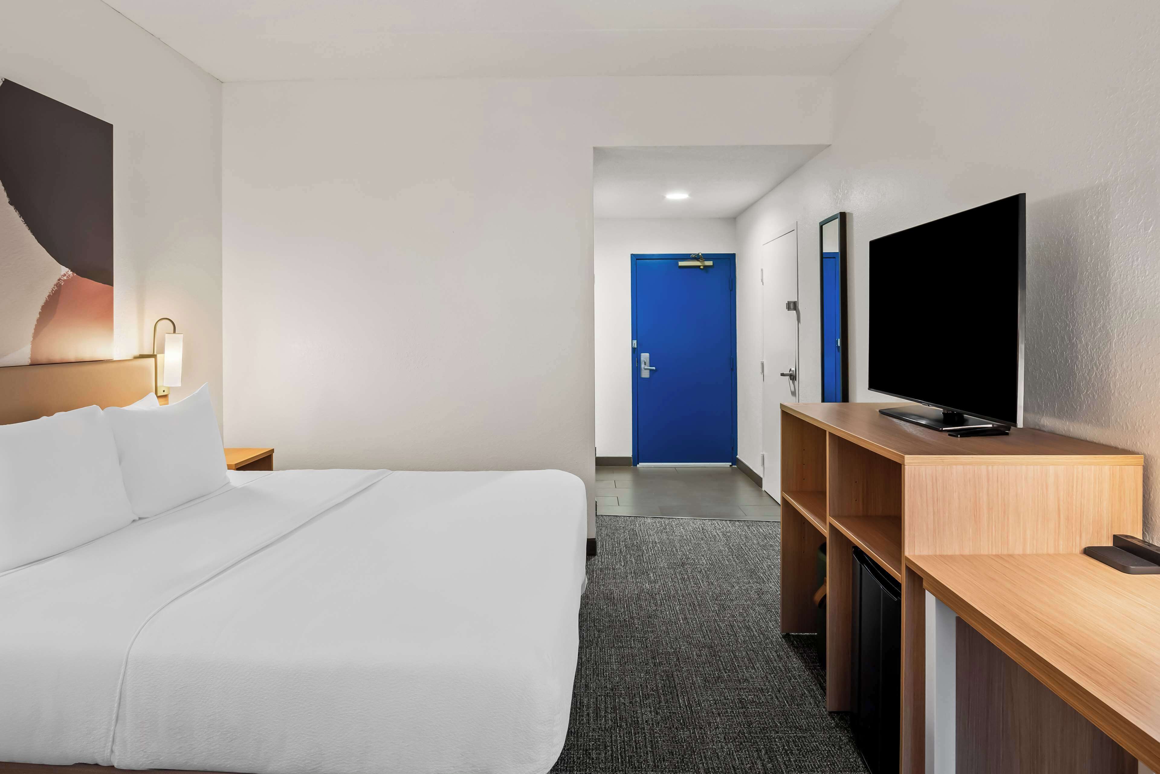 Habitación de hotel con cama, escritorio con televisor, puerta azul y mesita de noche con lámpara.