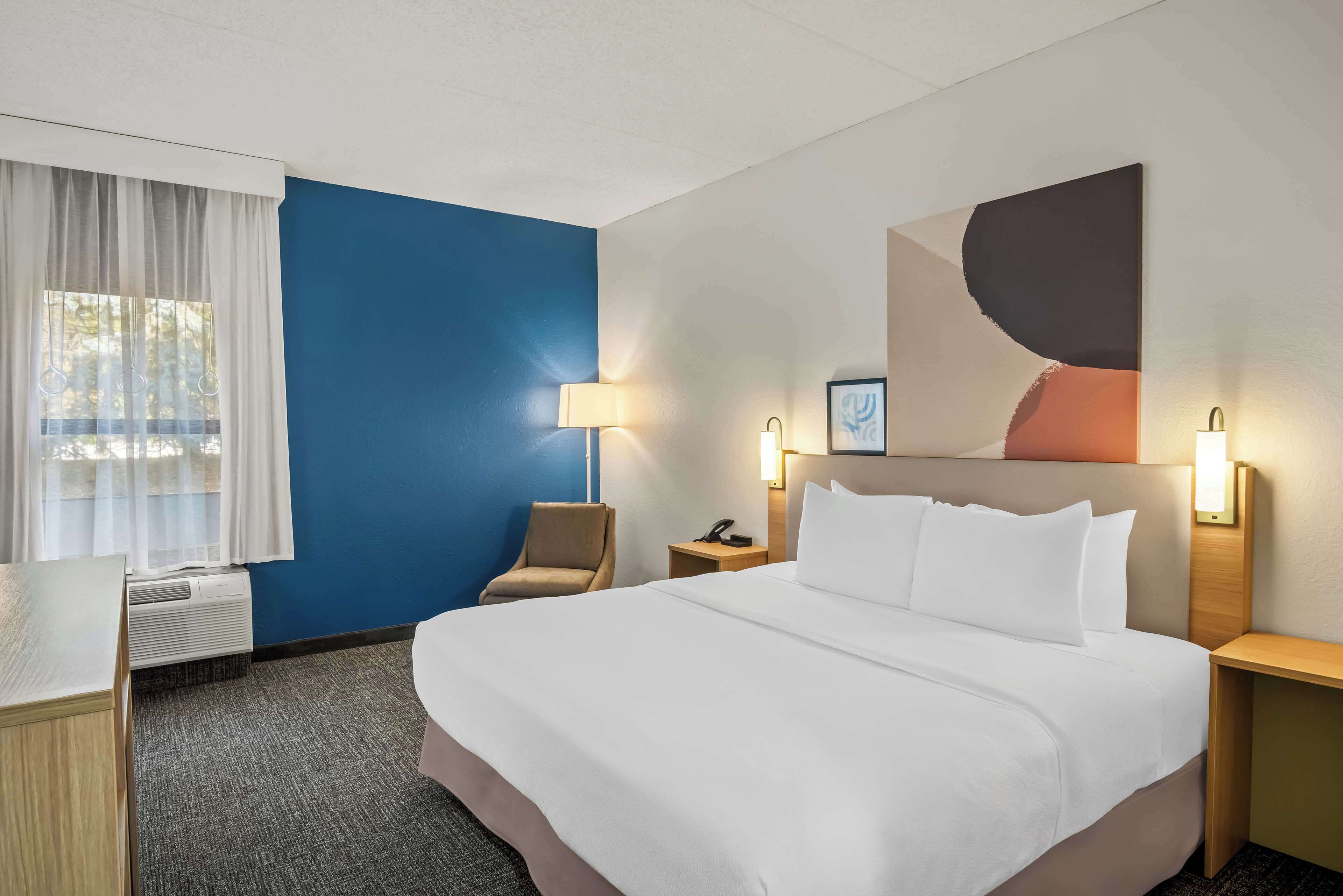 A hotel room with a large bed, a desk, a chair, a window with curtains, and a wall-mounted artwork.