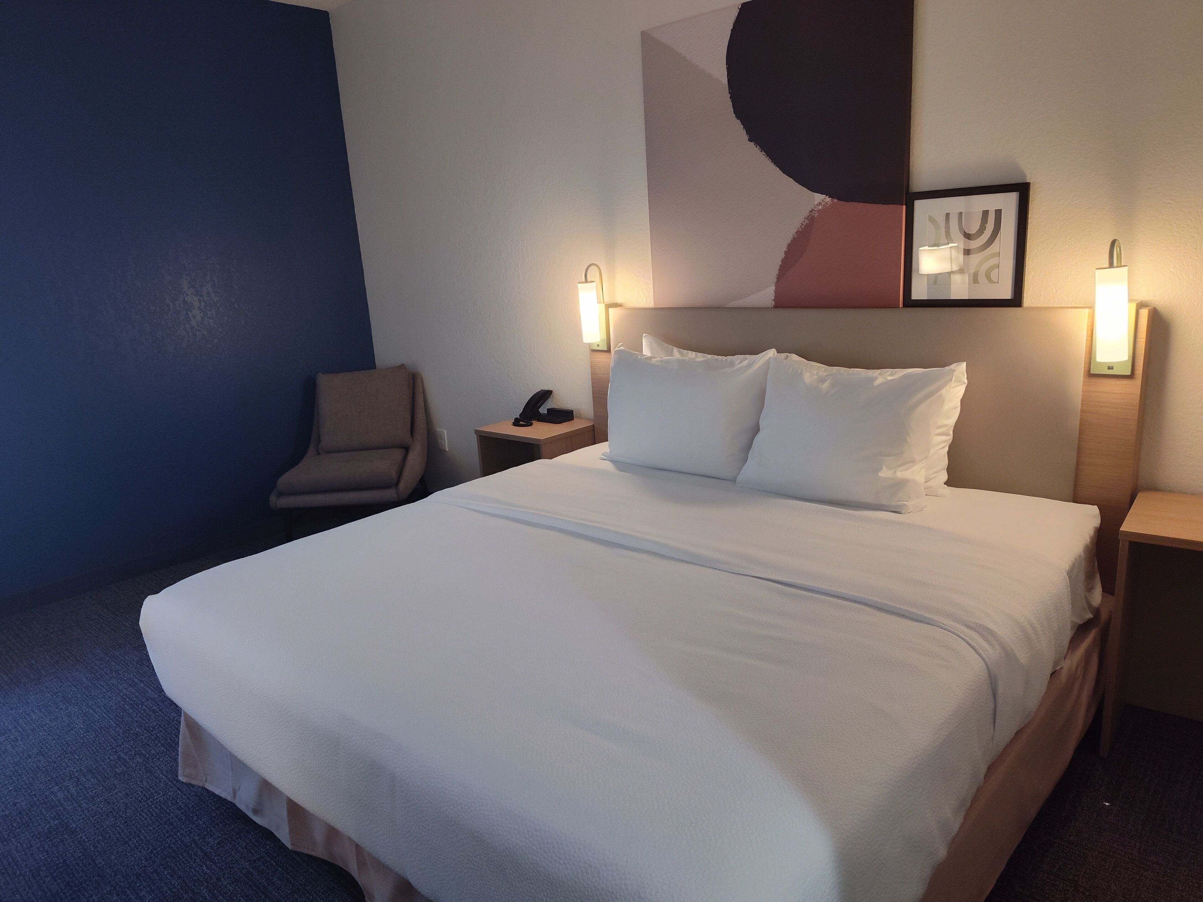 Una habitación de hotel con una cama grande, una silla, una mesita de noche y una lámpara fijada a la pared.