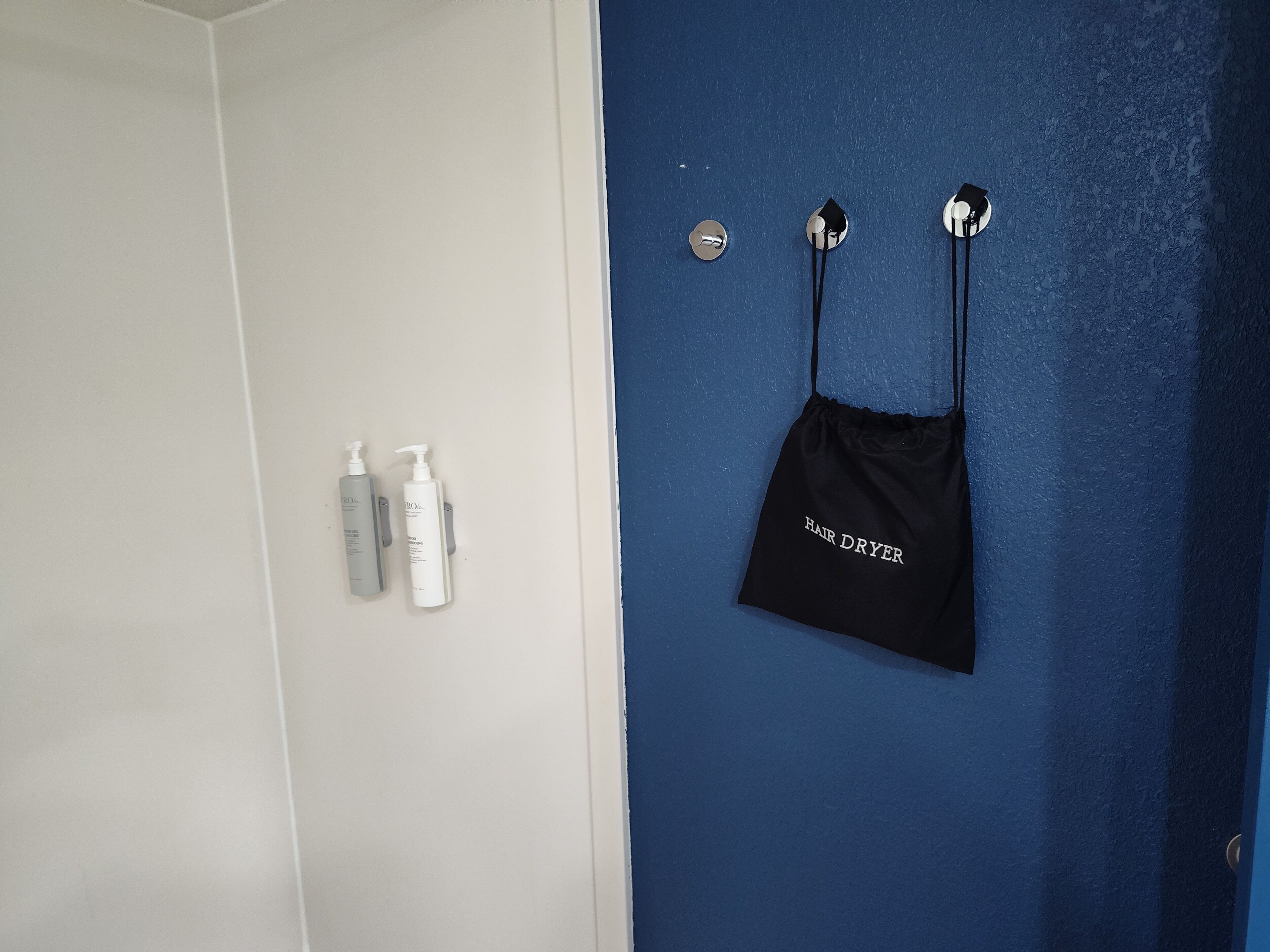 A bathroom with a white door and a blue wall. There are two bottles of hair care products on the wall and a black bag labeled "HAIR DRYER" hanging on the wall.