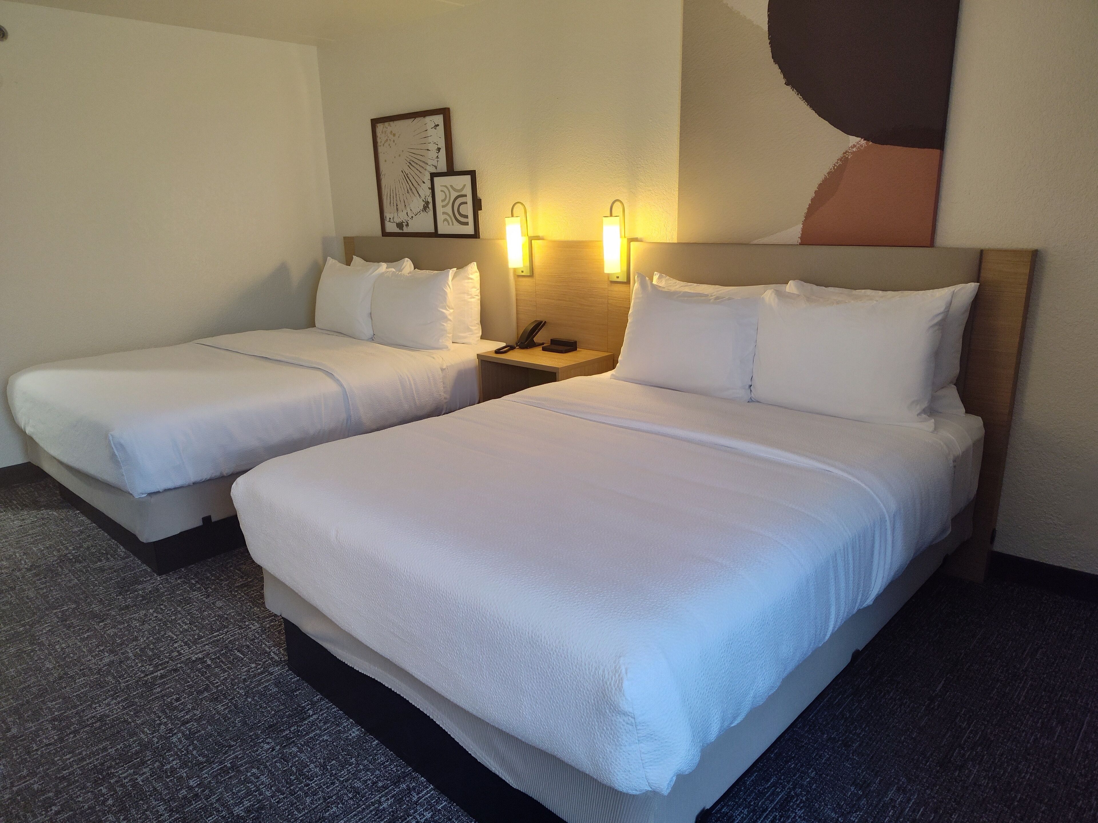A hotel room with two beds, a nightstand with a phone, and wall-mounted lights.