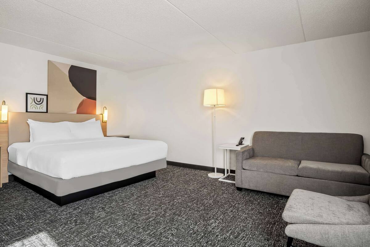 Ett hotellrum med en säng, en grå soffa, ett litet bord och en golvlampa.