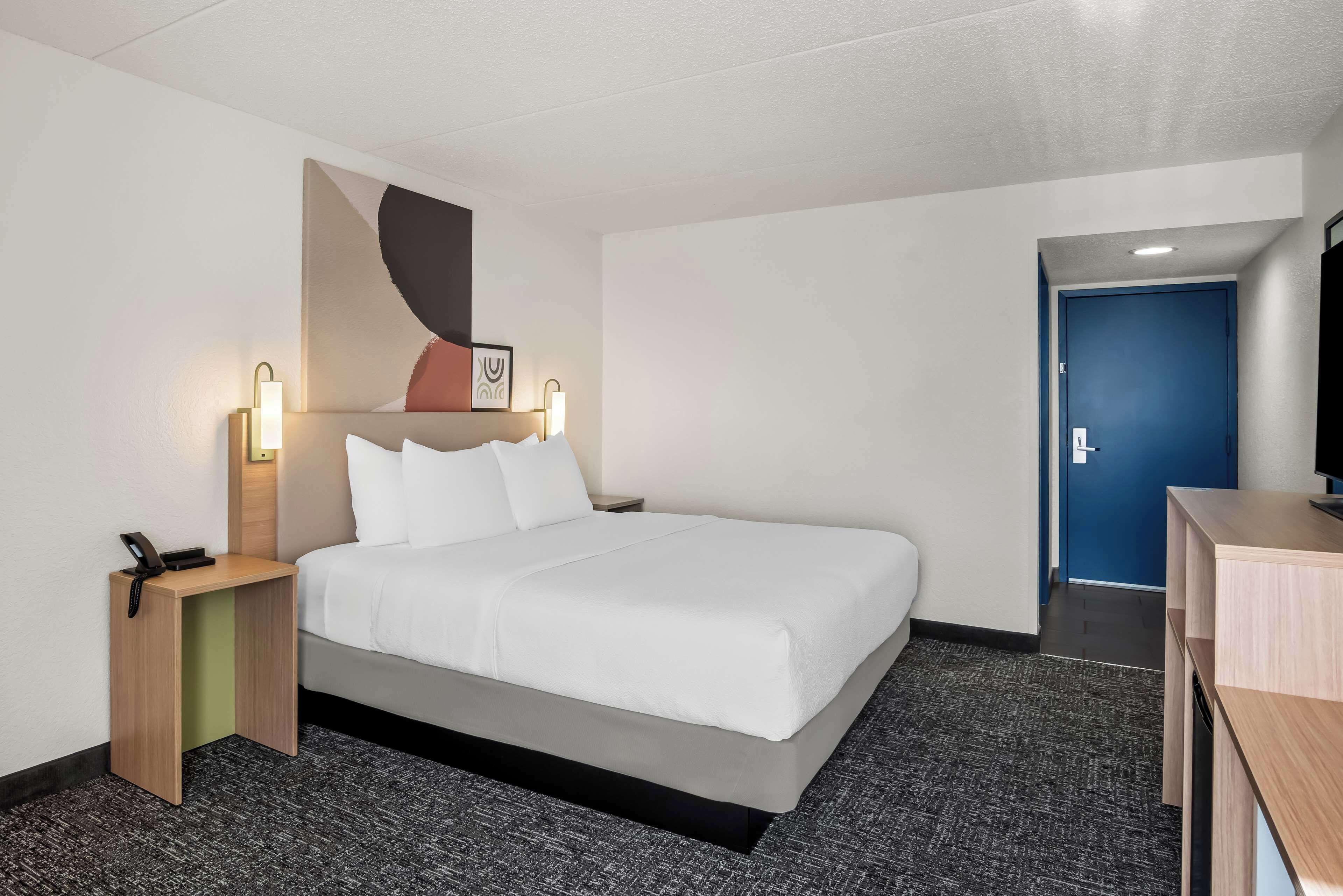 A hotel room with a bed, bedside table with a phone, wall-mounted light, and a door to another room.