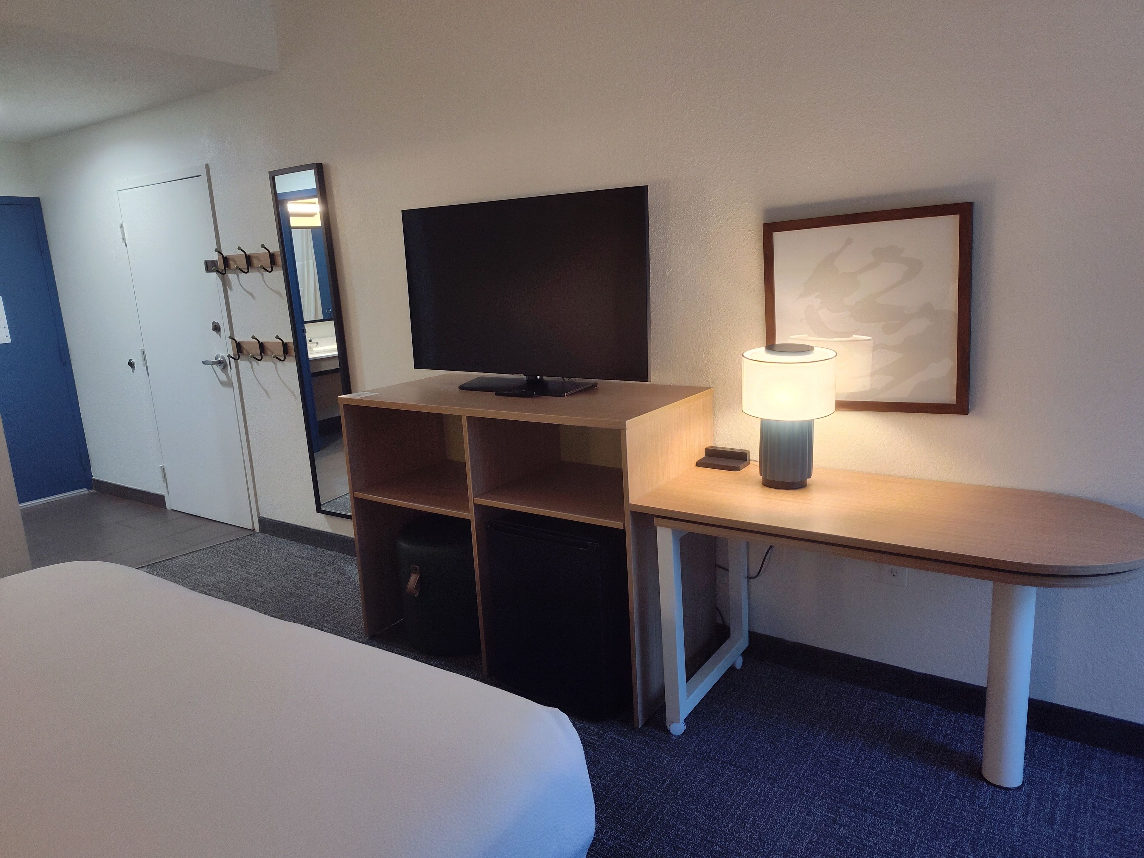 A hotel room with a bed, a desk with a television, a lamp, and a framed picture on the wall.