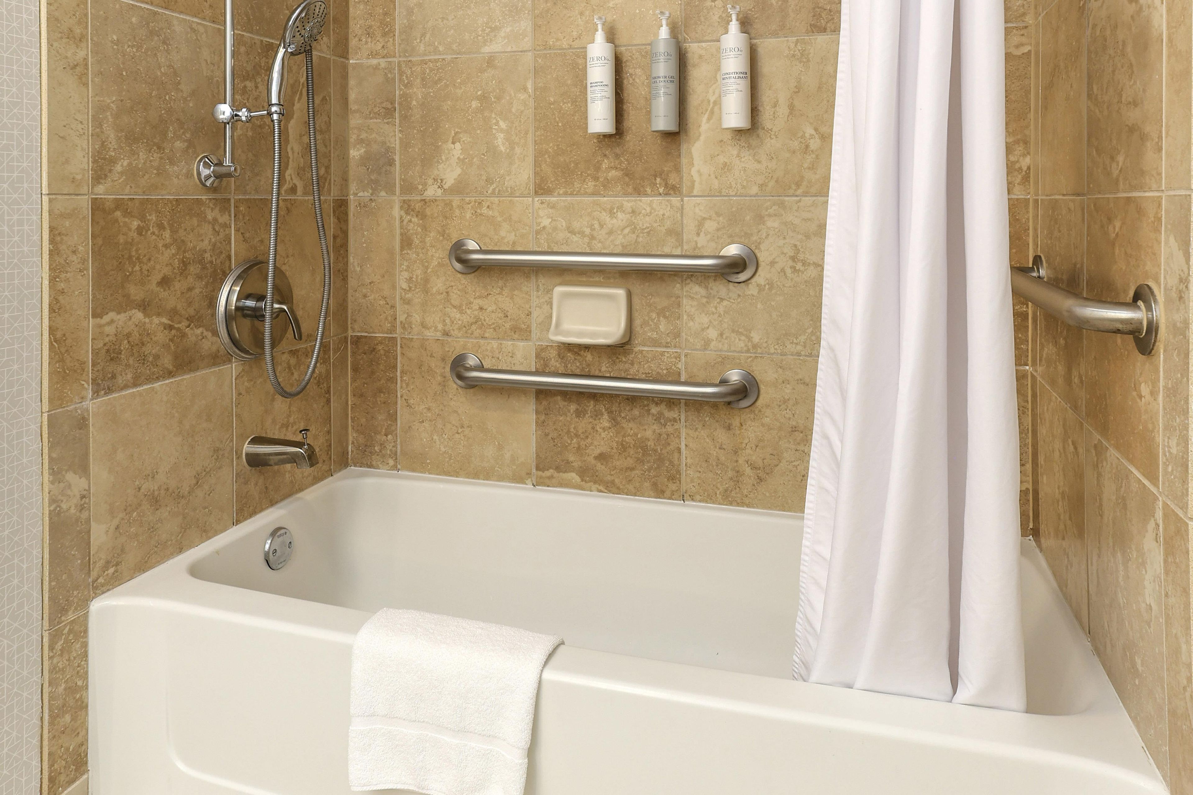 A bathroom with a bathtub, shower, grab bars, a towel, and toiletries.