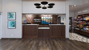 Zona de recepción de hotel con un mostrador de madera, un cartel que dice 'Welcome to Hampton', un mural con vistas a la ciudad y un bar de aperitivos con diferentes productos.