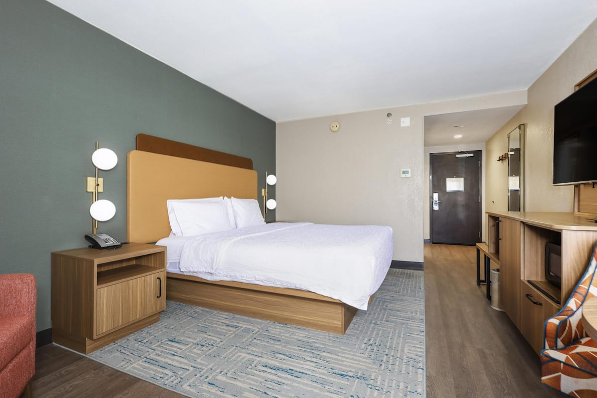 A hotel room with a bed, bedside table with a lamp, a TV, a desk with a chair, and a mirror.