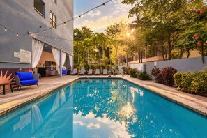 Una piscina con cielo iluminado por el sol, sillas de descanso y un edificio con porche cubierto.