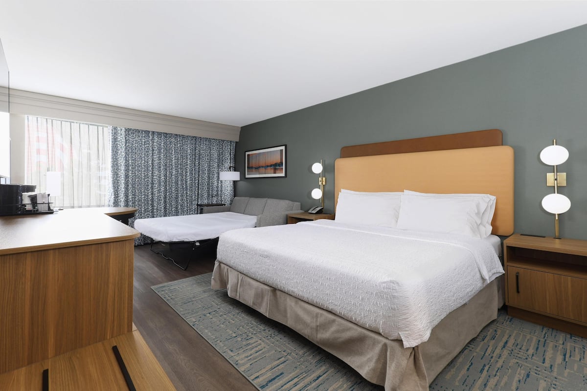 A hotel room with a large bed, a desk, a chair, a sofa, and a window with curtains.