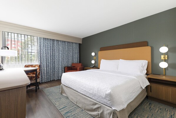 A hotel room with a bed, desk, chair, and a view of the outdoors.