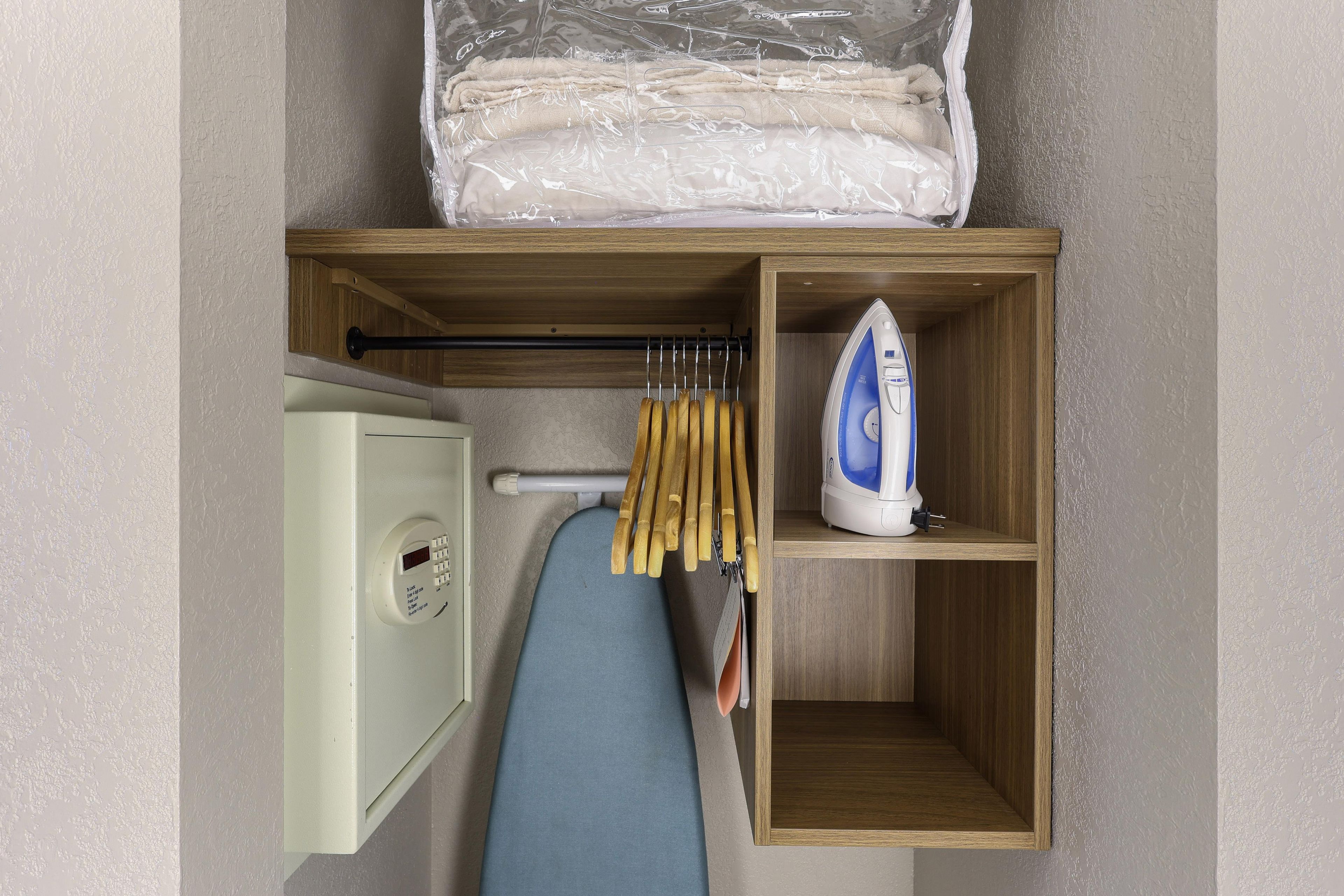 A compact storage unit with a hanging rack for clothes, an iron, and a small appliance, likely a dryer or washer, in a room with textured walls.