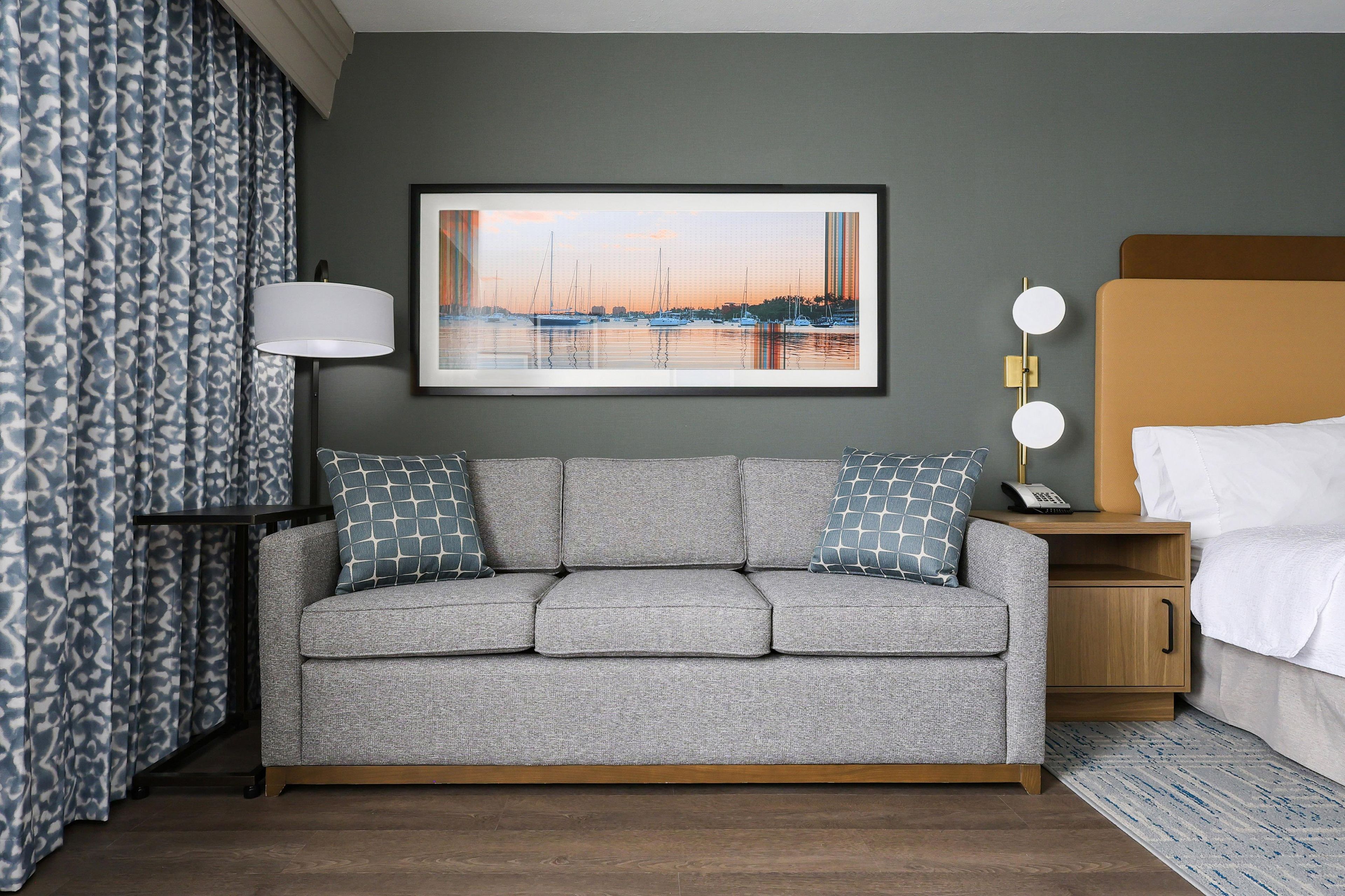 A hotel room with a grey sofa, a framed picture of a marina, a bedside table with a lamp, and a bed with white linens.
