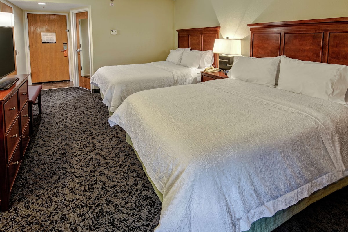 A hotel room with two beds, a wooden headboard, a bedside table with a lamp, and a carpeted floor.