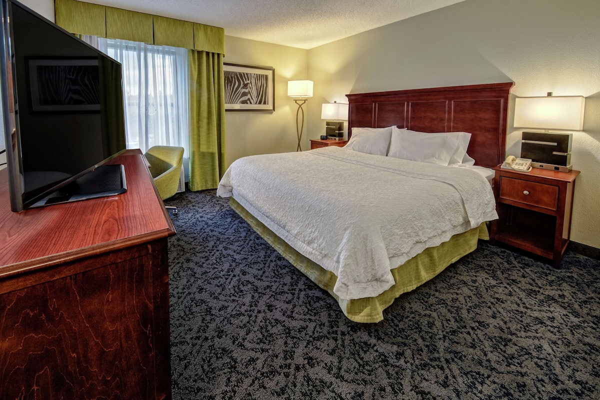 A hotel room with a large bed, a desk with a flat-screen TV, a chair, and a nightstand with a lamp and phone.