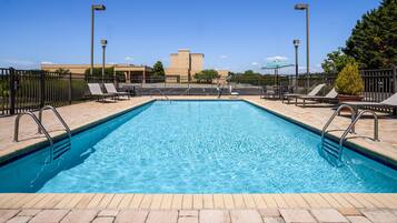 Una piscina rectangular con barandillas metálicas, rodeada de sillas de descanso y sombrillas, bajo un cielo despejado y con edificios al fondo.