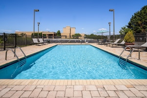Una piscina rectangular con barandillas metálicas, rodeada de sillas de descanso y sombrillas, bajo un cielo despejado y con edificios al fondo.