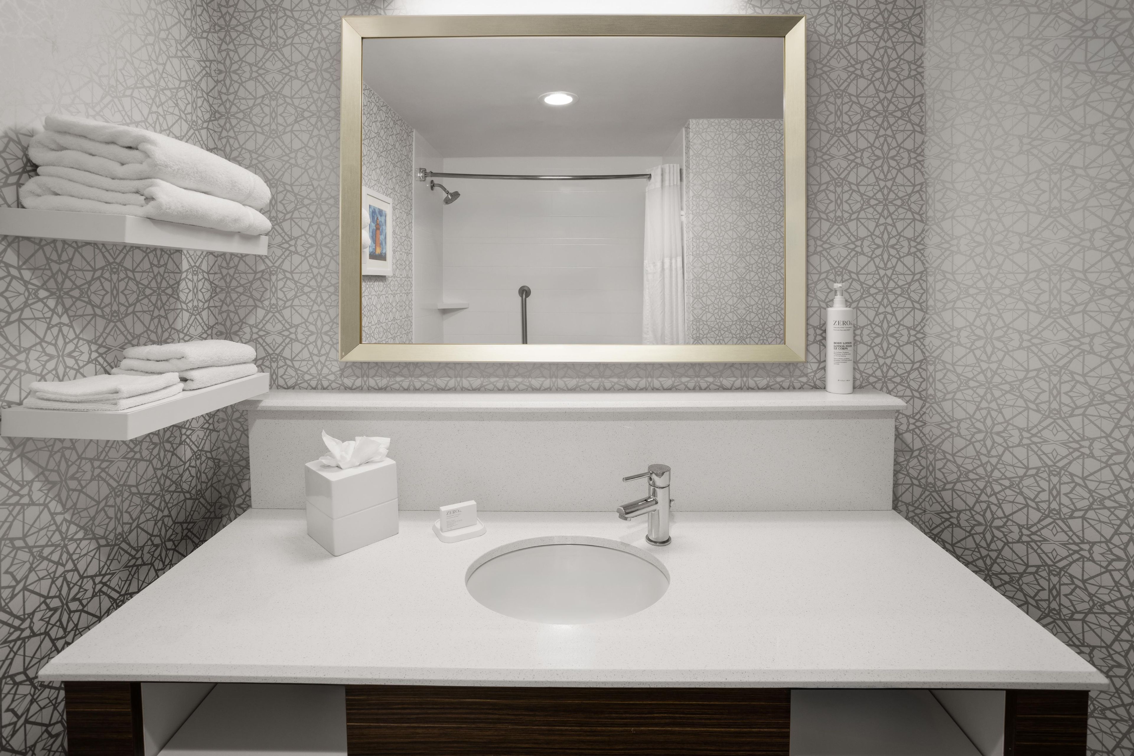 Une salle de bain avec un lavabo blanc, un miroir, un porte-serviettes et une étagère où sont pliées des serviettes.