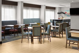 Un comedor de hotel con varias mesas y sillas, un televisor de pantalla plana montado en la pared y ventanas amplias con cortinas.