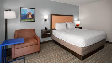 A hotel room with a bed, a chair, a desk, and a nightstand with a lamp.