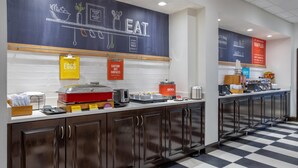 Ein sauberer, moderner Küchenbereich mit Schachbrettmuster-Boden, einer Tafel mit Zeichnungen zum Thema Essen und verschiedenen Verpflegungsstationen, darunter eine Eierstation und eine Haferflockenstation.