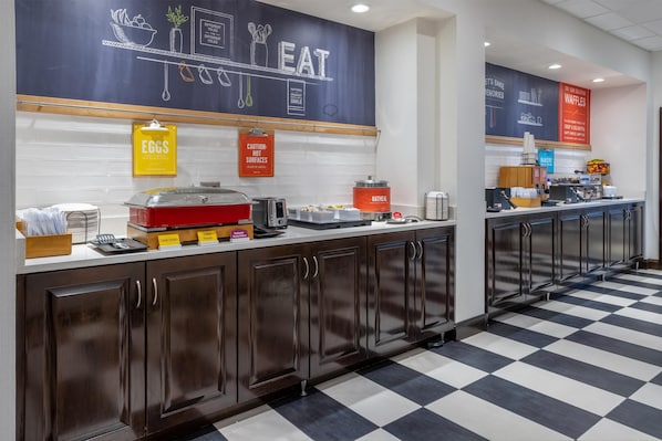 A clean, modern kitchen area with a checkered floor, a chalkboard with food-related drawings, and various food service stations including an egg station and an oatmeal station. - Hampton Inn Dover (Dover)