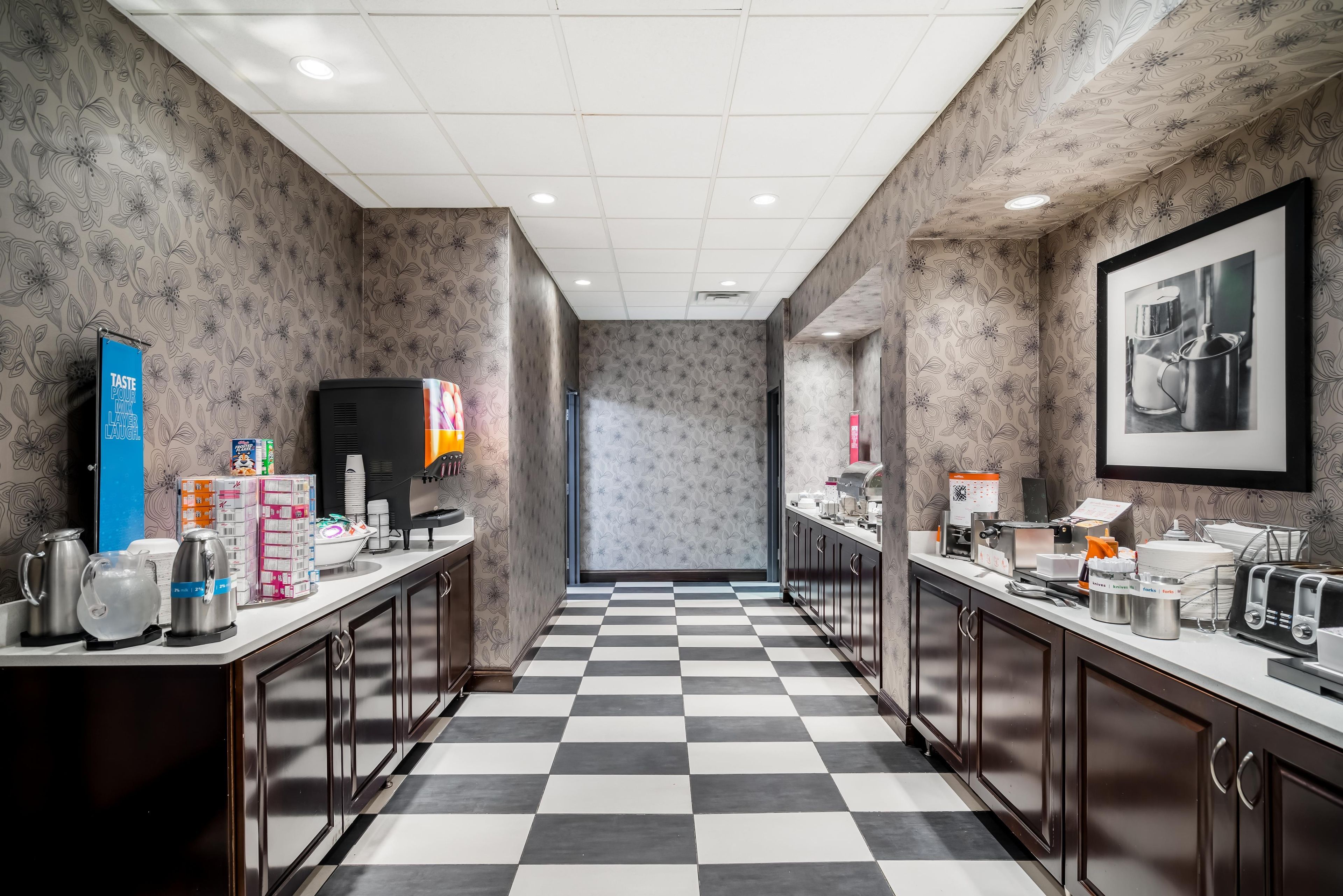 A hotel kitchen with checkered flooring, stainless steel appliances, and a framed photograph on the wall.