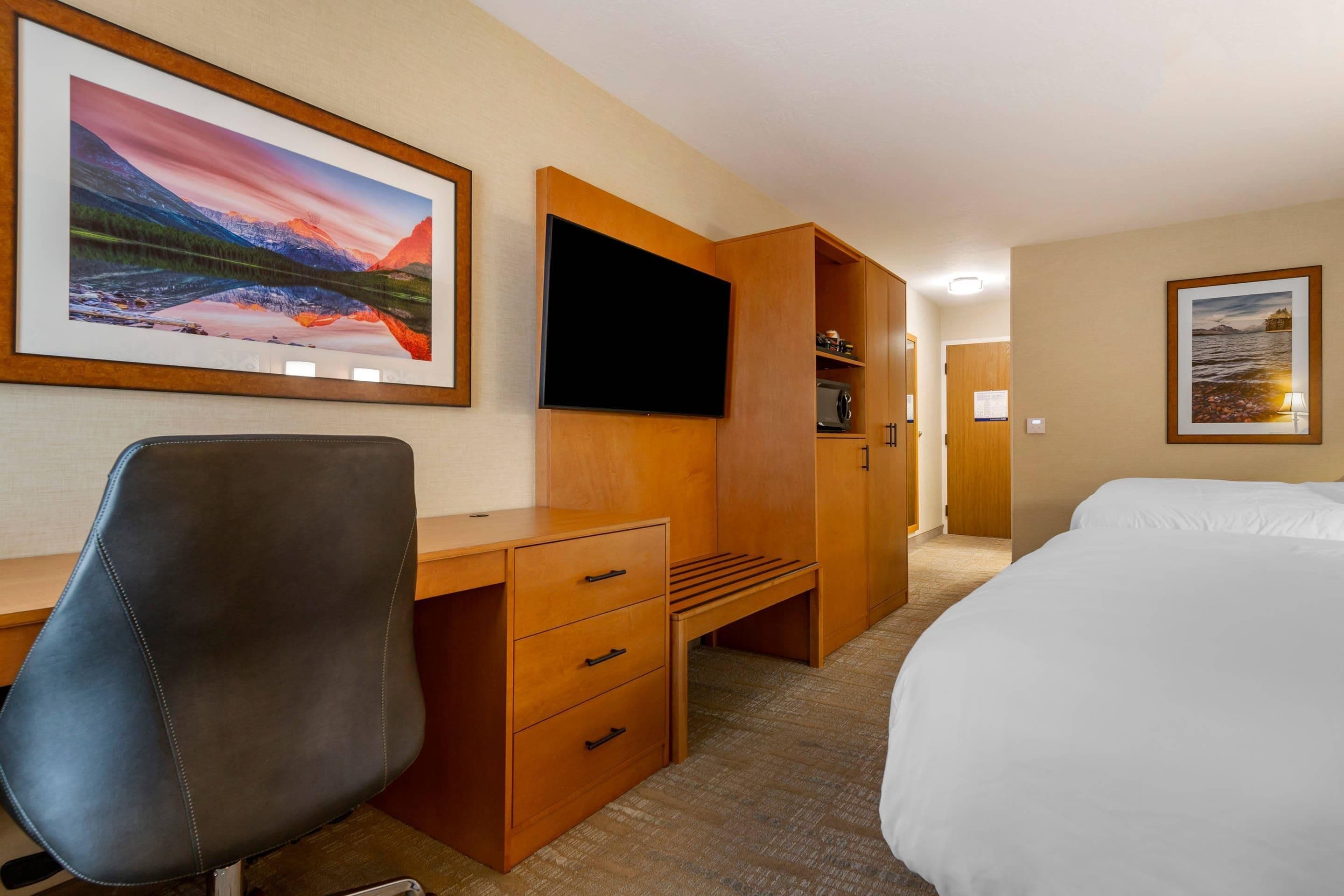 Una habitación de hotel con una cama, un escritorio con una silla, un televisor y obras de arte en las paredes.